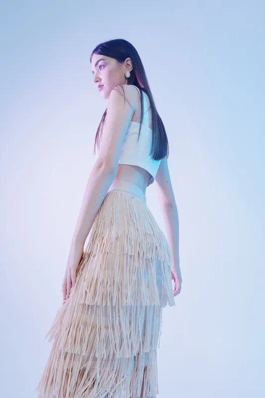 Young woman in a stylish fringe skirt and white crop top poses sideways in soft studio lighting. Concept of woman beauty fashion with elegant and modern style.