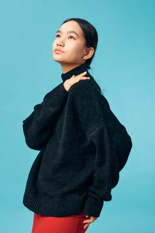 Young woman in black sweater with thoughtful expression, looking up against blue background. Casual fashion, portrait, stylish outfit, female model with natural makeup and long hair pulled back.