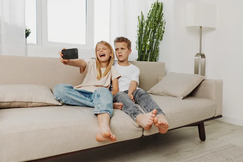 Two children sitting barefoot on a beige sofa taking a selfie with a smartphone in a bright living room. The girl is laughing while the boy looks serious. Casual home moment with siblings.