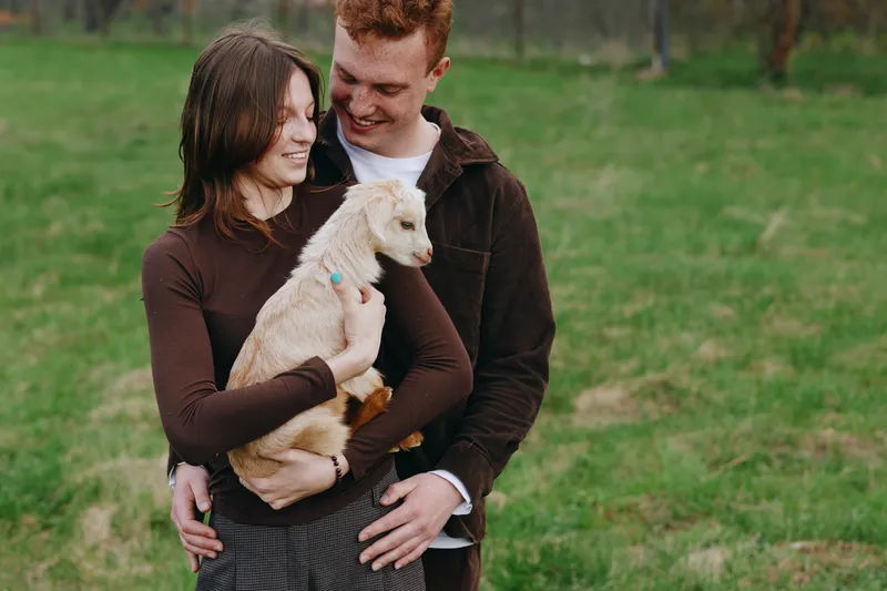A couple outdoors in a grassy countryside hold a fluffy goat kid, smiling and affectionate. Warm natural light enhances the rustic scene, conveying family, pets, and gentle companionship in nature.