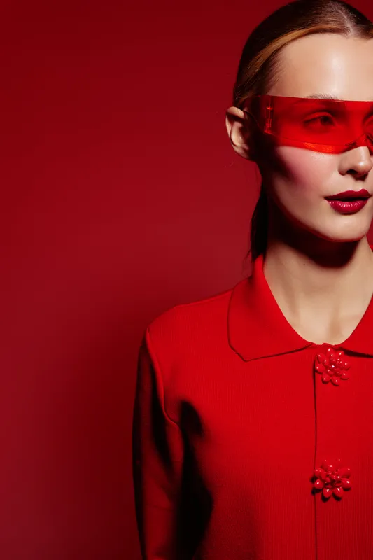 Fashion model in red sweater and red translucent visor poses against red background. Concept of modern style, bold color, minimalism, and futuristic fashion portrait with strong contrast.