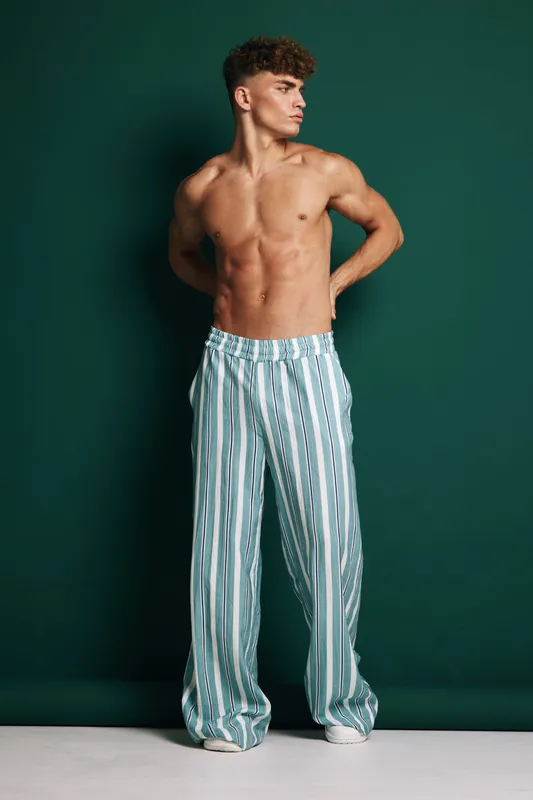 Athletic young man standing shirtless with hands behind back wearing blue striped pants and white shoes on clean background, displaying strong toned muscles in casual pose.