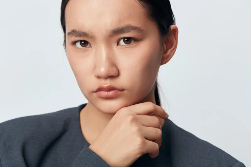Young woman with serious expression, hand on chin in thoughtful pose against plain background. Closeup portrait showing focus, contemplation, and introspection in studio setting.
