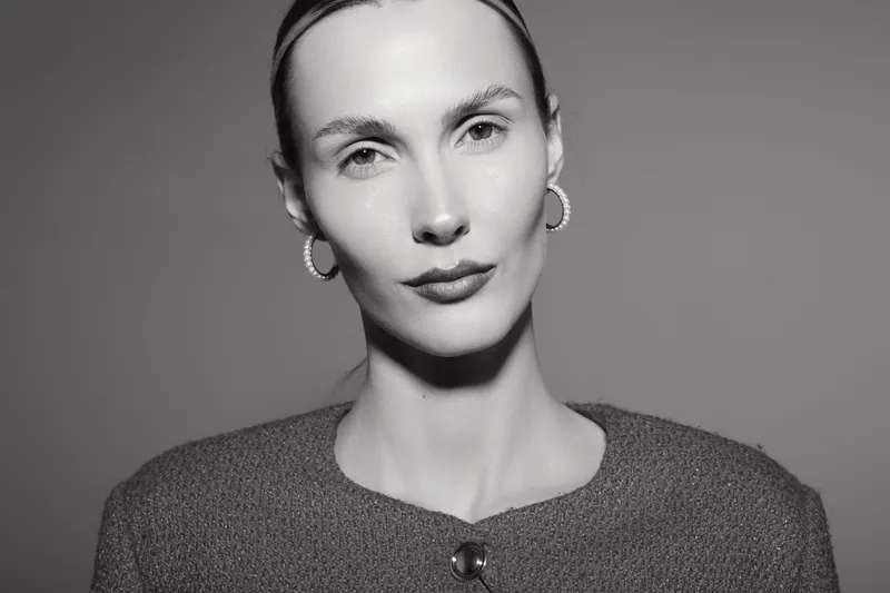 Portrait of a young Caucasian woman with pale skin and slicked-back hair wearing hoop earrings and a textured sweater. Neutral expression on a studio grey background, fashion concept.