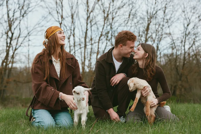 Three friends outdoors with dogs enjoying a relaxed moment in a grassy park, warm brown tones, casual outfits, natural light, candid interaction, and a sense of companionship