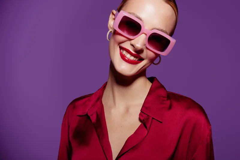 Smiling woman in pink sunglasses and red blouse poses against purple background. Bright makeup and confident expression create a stylish and modern fashion portrait concept.