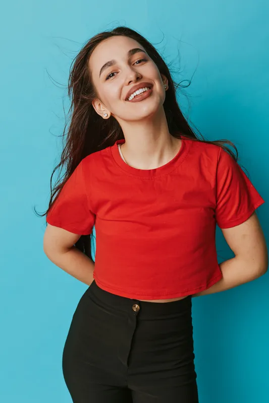Young woman smiling and posing with hands behind back wearing red shirt and black pants against blue background. Happy female model with long hair in casual outfit looking confident.