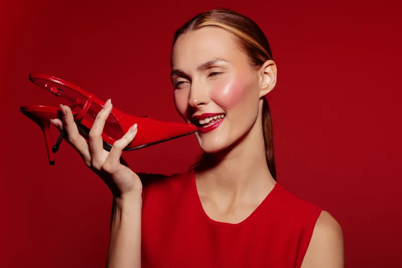 Smiling woman in red dress holds red high heel shoe near face on red background. Concept of fashion, style, beauty, and bold color contrast with playful mood.