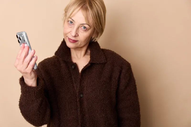 An elderly woman holds a smartphone in a studio portrait, beige background, thoughtful expression, communication and technology, casual home style.