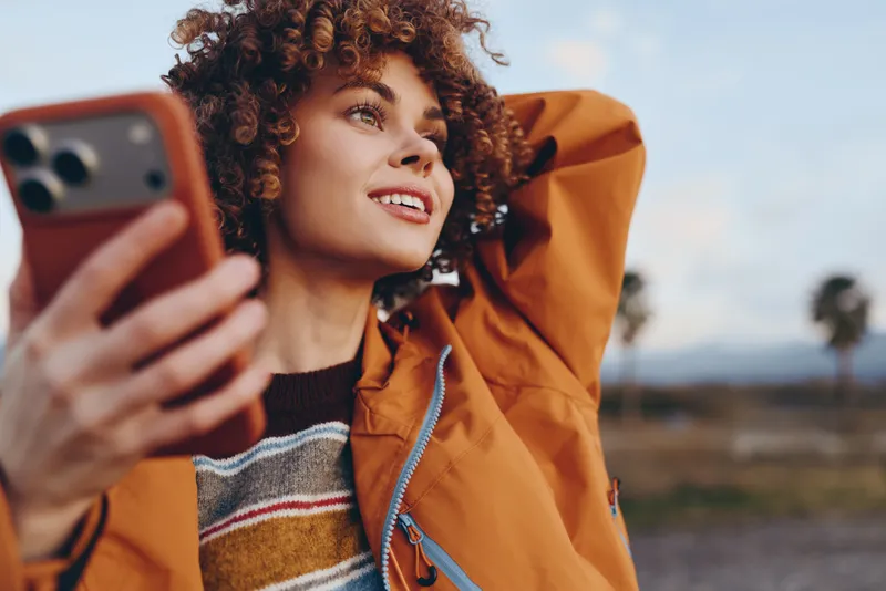 Woman lifestyle selfie phone outdoors curly jacket orange portrait, smiling woman holds smartphone near face, relaxed outdoor moment on countryside field with soft sky light