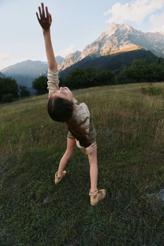 A young woman dances in a grassy meadow, arms raised toward the sky, mountains in the background, conveying freedom, vitality, and a carefree outdoor lifestyle.