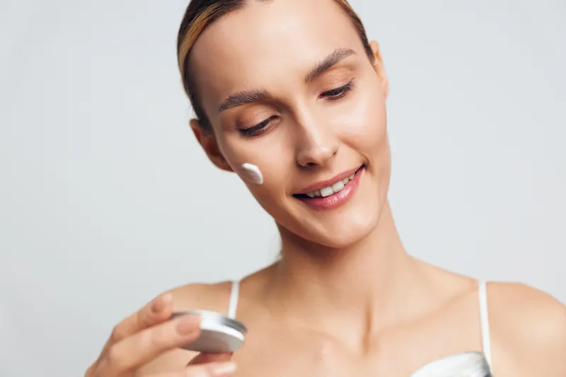 Young woman with brunette hair applies white face cream on her cheek while smiling gently against a plain light background in a skincare routine moment.