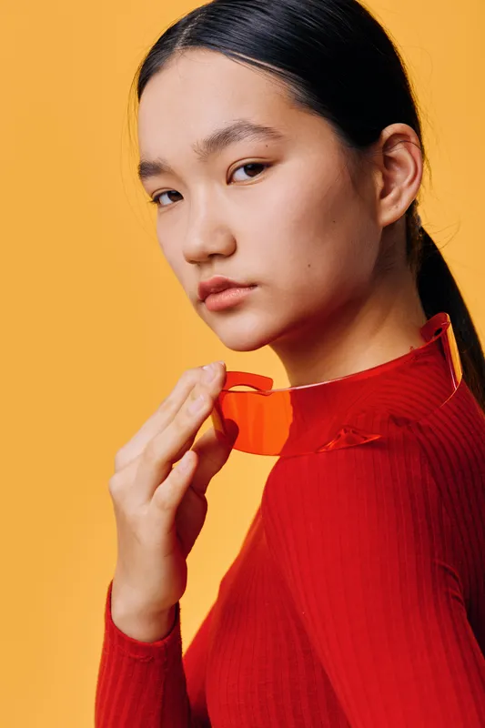 Young woman portrait with long dark hair wearing red turtleneck sweater and holding red glasses near face on orange background for fashion and style concept.