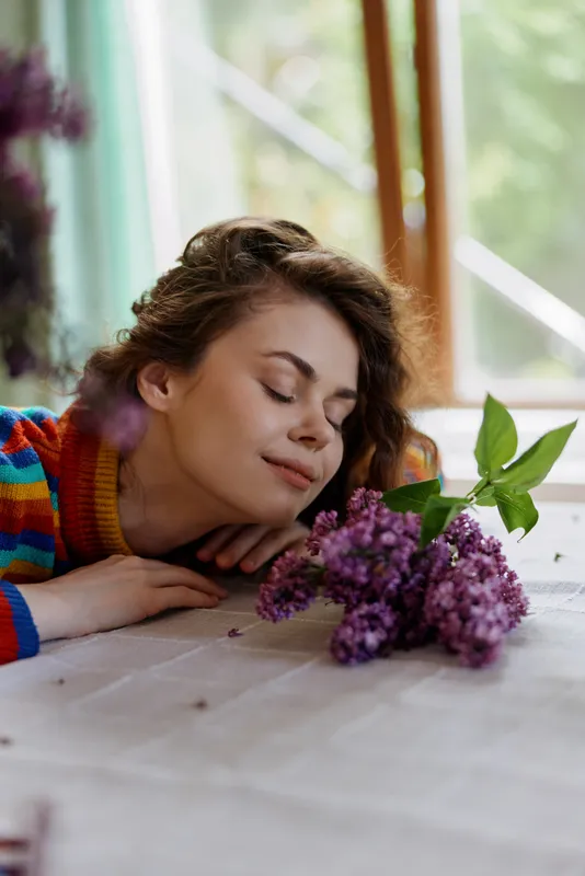 A young woman rests her head on a white wooden table beside a lilac bouquet in a bright cozy home, eyes closed and a serene smile, conveying peaceful relaxation indoors near a window.