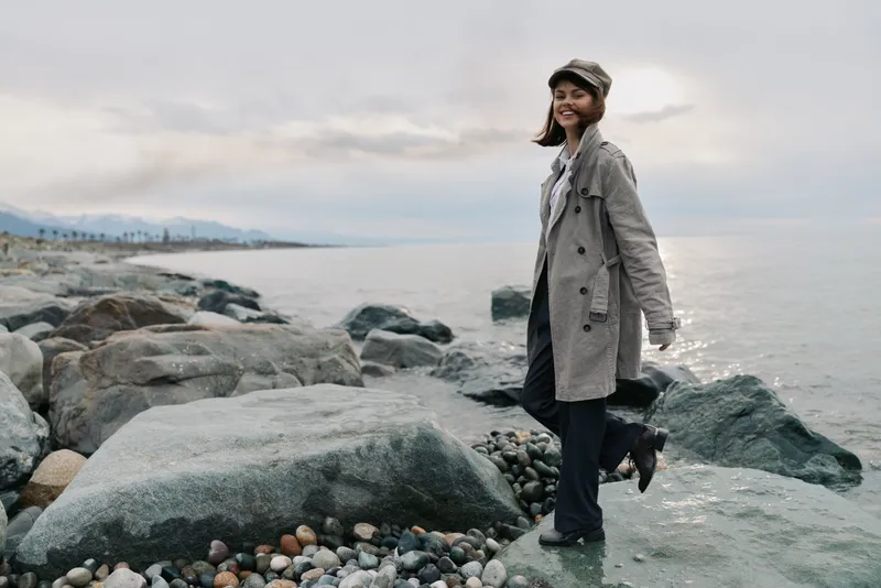 Young woman in a plaid coat and cap stands on rocky shore near calm sea under cloudy sky. Casual fashion and outdoor lifestyle concept with natural landscape background.
