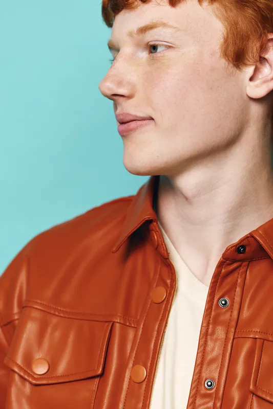 Young man with red hair wearing brown leather jacket and white shirt looking sideways on blue background. Casual style profile portrait with natural light and soft expression.