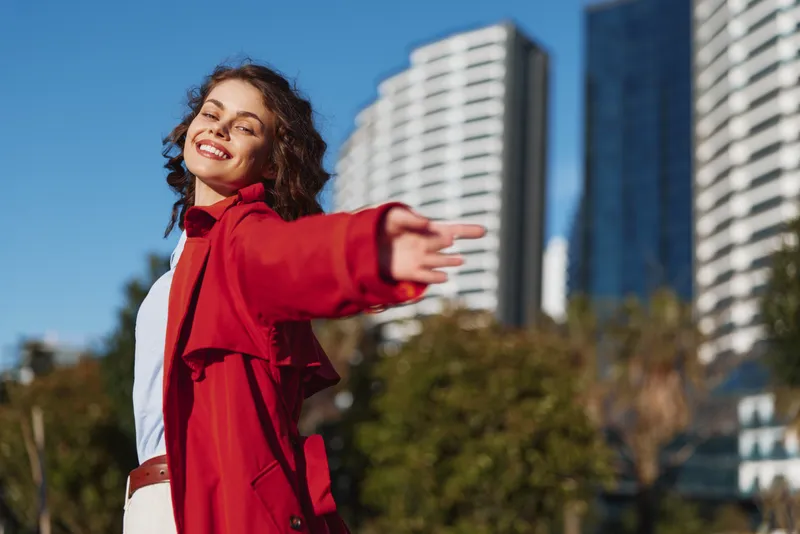 Young woman with curly hair wearing a red jacket smiles joyfully while extending her arm outdoors in an urban environment on a sunny day.