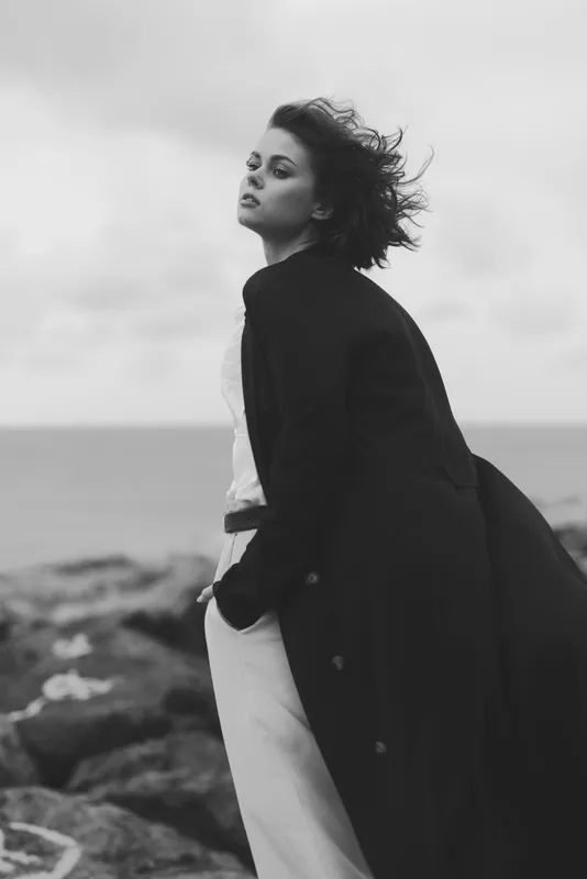 Moody black and white photo of a stylish woman on a windy shoreline wearing a long coat, flowing hair, and confident stance, capturing fashion mood and timeless coastal elegance.