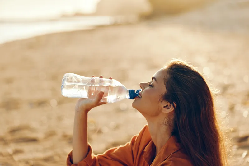 Woman drinking water outdoors in sunlight during summer, health and hydration concept, mature woman in orange top in dry desert landscape, calm expression, warm natural light day