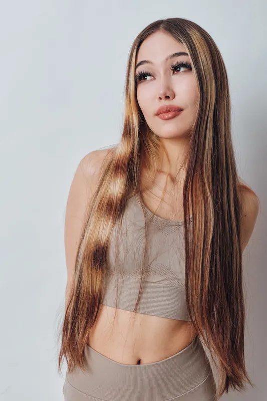 Young woman with long straight hair and neutral makeup wearing beige crop top and leggings standing against light background in studio, looking sideways with calm expression.