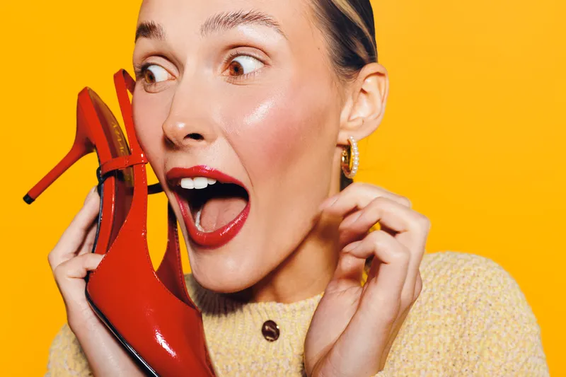 Surprised woman with red lipstick holds red high heel shoe near face on bright yellow background. Concept for fashion, style, beauty, and shopping excitement or discovery.