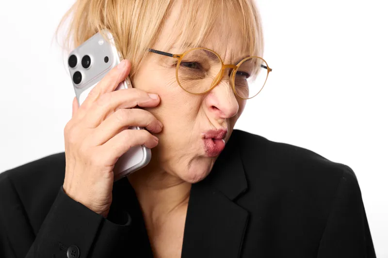 Blonde business woman with glasses holds phone to ear, lips pursed, showing stress and surprise, studio white background, workplace communication for an urgent call