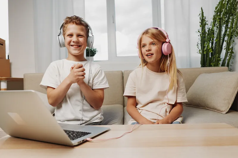 Two cheerful children wearing headphones sit on a couch enjoying music from a laptop in a bright living room. Concept of leisure, technology, and happy childhood moments.