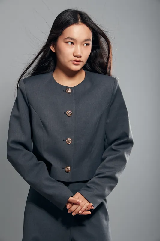 Young woman with long dark hair wearing a gray buttoned jacket and matching pants posing against a neutral gray background, looking sideways with clasped hands.
