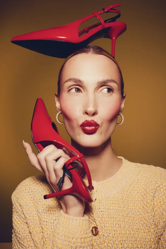 Stylish woman with red high heels poses playfully against yellow background. She wears hoop earrings and a textured sweater, showcasing fashion and bold accessories concept.