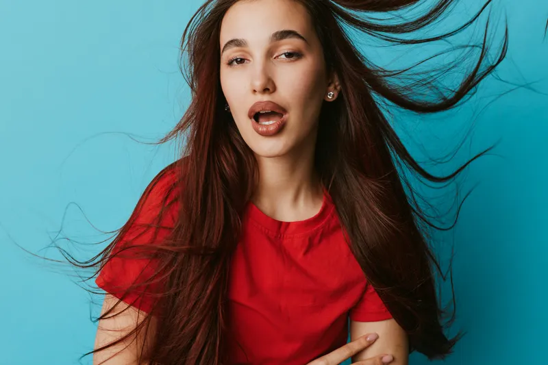 Woman with long hair and red shirt posing with wind blowing hair on blue background. Female model with expressive face and casual style in studio portrait photography.
