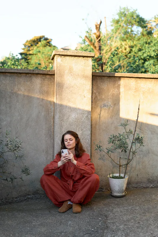 Casual woman in a rust jumpsuit sits on a courtyard ledge, looking at her phone. Warm afternoon light highlights a relaxed lifestyle moment amid a weathered wall and a potted plant