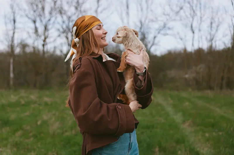 A joyful woman outdoors holds a fluffy animal close, smiling as they share a tender moment in a grassy field. A lifestyle portrait conveying warmth, connection, and affection between human and pet.