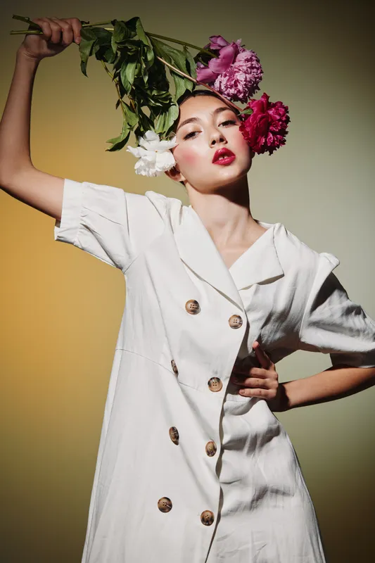 Fashionable young woman in white dress posing with vibrant peony flowers held overhead against neutral background. Concept of beauty, creative style, and floral fashion.