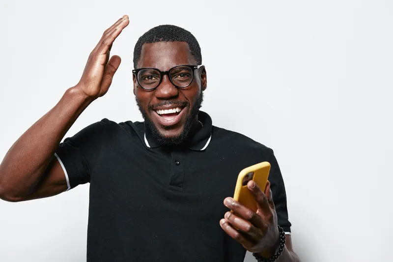 African American man with dark skin and glasses smiling and holding yellow smartphone in black polo shirt on white background. Happy male with beard using mobile phone for communication.
