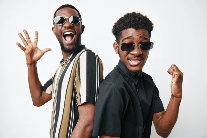 Two young African American men with dark skin wearing sunglasses and casual shirts posing back to back with happy and confident expressions on white background studio.