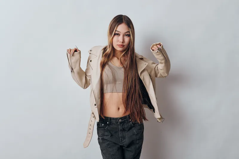 Young woman with long hair wearing beige jacket and crop top posing confidently with raised hands on white background. Casual style, fashionable outfit with neutral tones in studio.