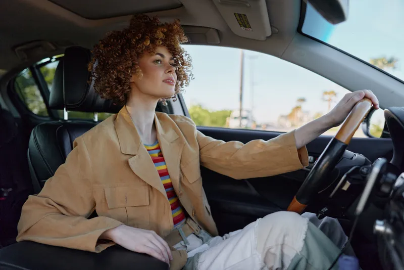 A stylish woman driver sits behind the wheel in a tan blazer and striped top, looking confident yet relaxed. Warm retro film tones enhance the mood of everyday urban travel.