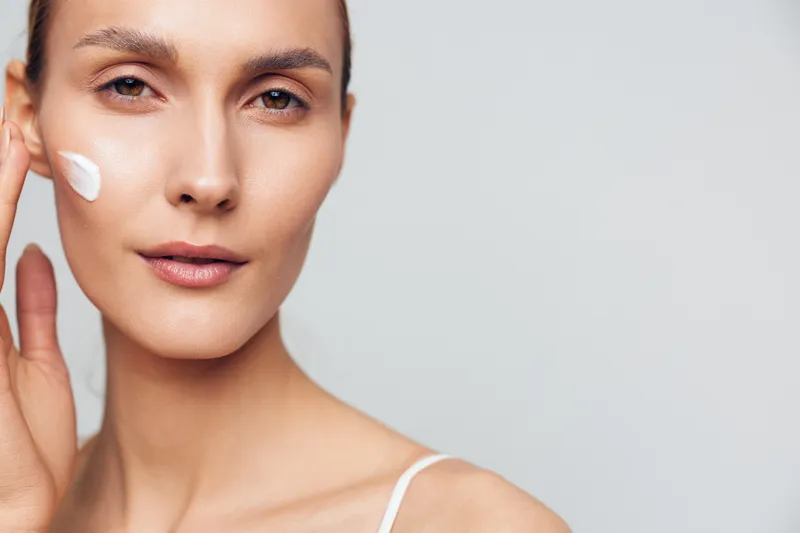 Young woman applies cream to her cheek in a close up portrait, calm expression, soft lighting, skincare moment, clean minimal background
