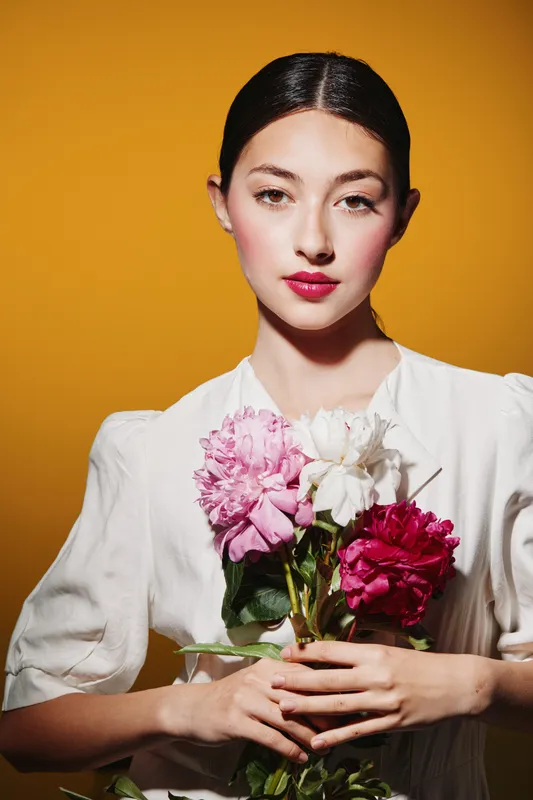 Elegant young woman with natural makeup wears a white blouse and holds vibrant pink peonies against bright yellow background. Concept of beauty, fashion, sophistication, and floral inspiration.