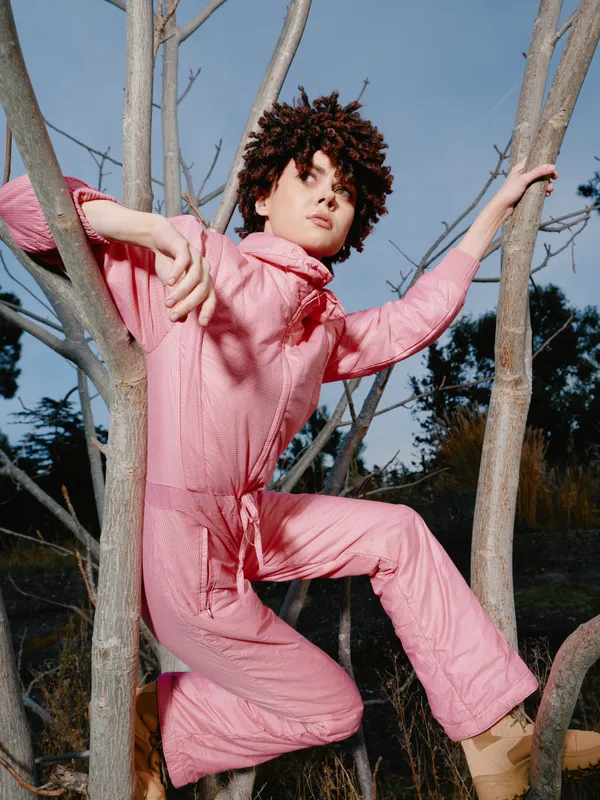 Person climbing tree in a pink jumpsuit outdoors, showcasing bold fashion and athletic energy with dynamic pose, sky and branches in view for a vibrant nature portrait.