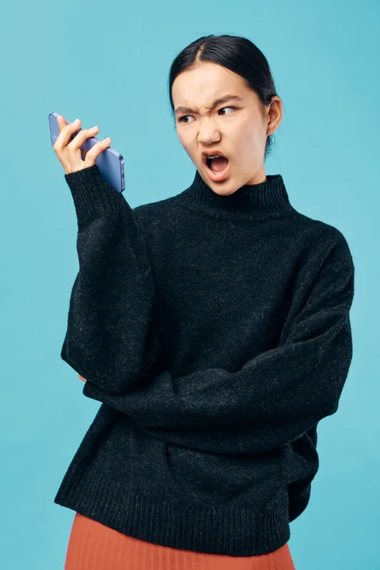 Angry woman in black sweater holding smartphone with furious expression on blue background. Emotion of frustration and upset reaction to phone call or message.