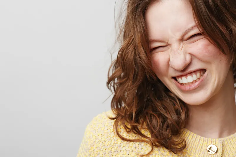 Close-up of a young woman with curly brown hair wearing a yellow sweater grimacing with eyes closed. Studio portrait on a light grey background with natural expression and casual style.