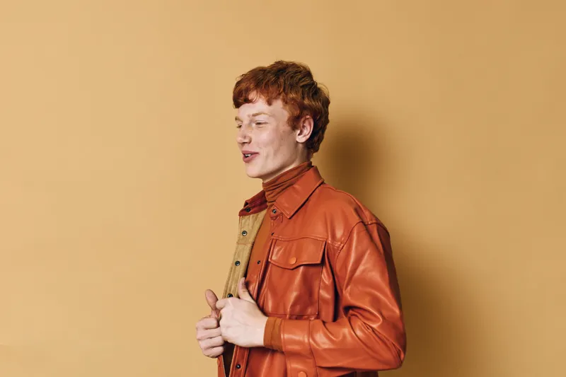 Young man with red hair wearing brown leather jacket and turtleneck standing against beige background looking sideways with confident expression and casual posture.