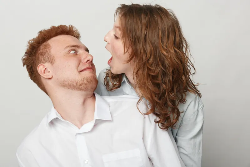 Curly woman playfully shouting at surprised man with red hair in white shirts on grey background. Concept of fun, communication, emotions, and relationship dynamics between young adults.