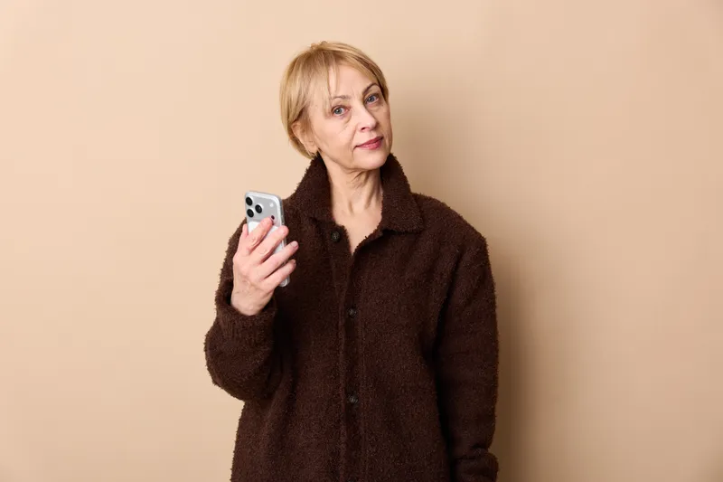 Elderly woman holding a smartphone, looking up as if checking messages, Studio portrait on a beige background, communication and technology theme, calm mood