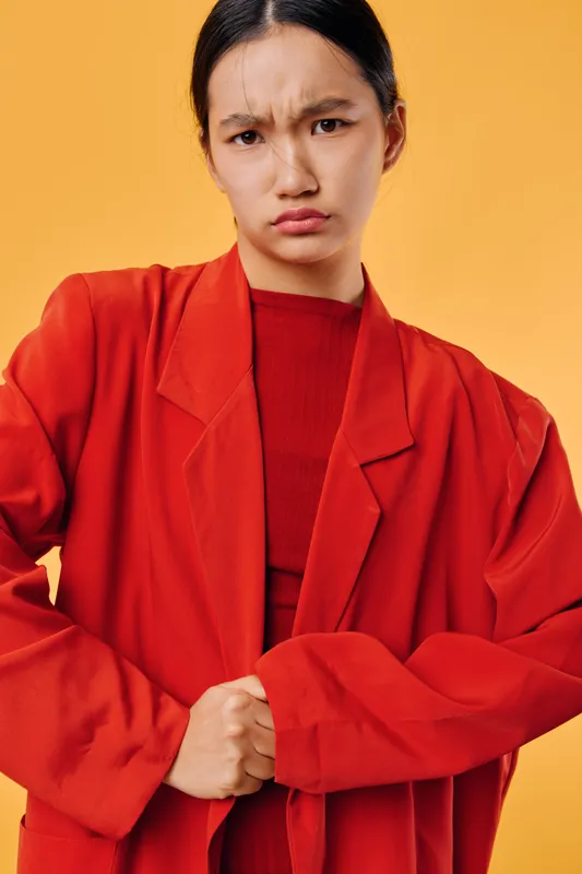 Young woman with serious facial expression wearing red jacket and sweater posing against yellow background. Portrait of confident model with natural look and casual style.