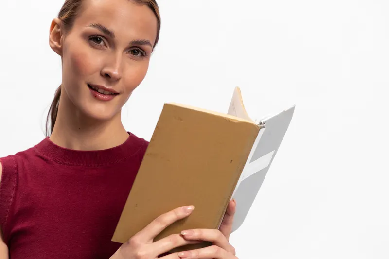 Young woman in red sweater holding an open book looks at the camera, isolated on white background. Concept of reading, education, and concentration.