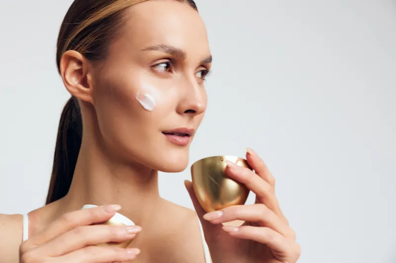 Young woman applies face cream on cheek holds gold jar with skincare product has smooth skin and natural makeup isolated on white background with focused expression.
