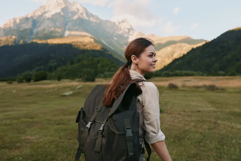 Young woman with a backpack hikes through a grassy valley with mountains in the background, looking ahead with a calm expression on a bright day in a wide open outdoor setting.