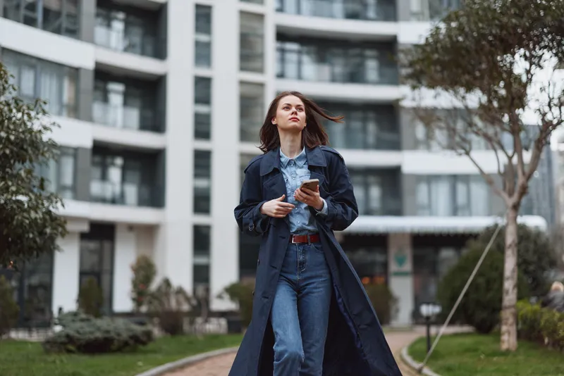 Young woman with long brown hair walks confidently through an urban park wearing a dark trench coat and denim jeans while holding a smartphone in her hand.